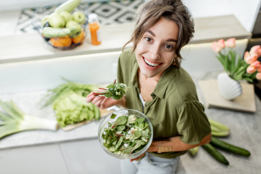 Portrait of a young and cheerful woman eating salad standing on the kitchen with food ingredients on the background, view from above. Vegetarianism, wellbeing and healthy lifestyle concept
