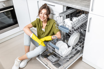 Portrait of a happy housewife sitting near the dishwasher with clean dishes on the kitchen at home....
