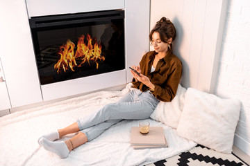 Young woman relaxing with a smart phone, sitting near the fireplace at the modern living room at...