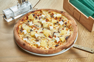 Appetizing baked pizza with white sauce, chicken, pineapple and mushrooms with a crispy crust on a wooden background. Restaurant table setting. Close up