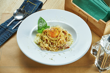 An appetizing portion of homemade Italian carbonara pasta(spaghetti) with egg yolk, bacon and parmesan in a white plate on a wooden background. Restaurant table setting