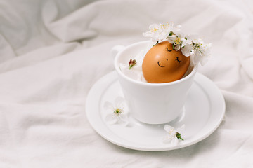 Funny Easter eggs with a wreath of flowers in a cup. Tender easter composition on white fabric background.