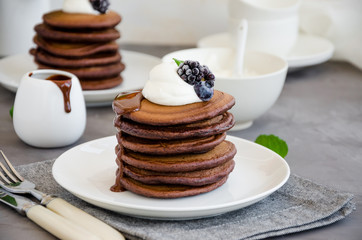 Stack of chocolate pancakes on a white plate with chocolate sauce, whipped cream, blueberries and blackberries on top of a concrete background. Delicious breakfast. Horizontal orientation. Copy space.