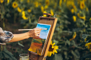 Close up hands of female artist holding brush and draws a picture of the field. Blurred background...