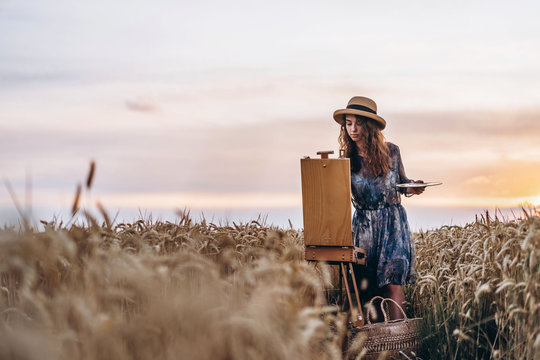Portrait of smiling female artist with curly hair in hat. Girl draws a picture of a landscape in a wheat field. Copy space