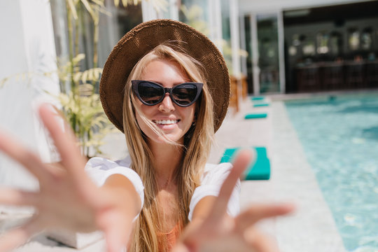 Glad young woman in hat and glamorous sunglasses making selfie at resort. Stunning female tourist with tanned skin chilling near pool.