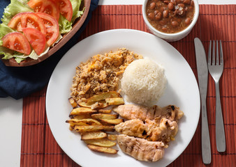 Dish of grilled chicken fillet with rice, beans, french fries and farofa on white plate. Top view.
