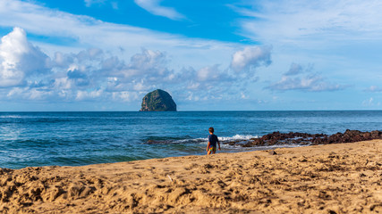 Fototapeta na wymiar Enfant jouant sur une plage de sable blanc devant une mer bleu d'azur avec un rocher en forme de diamant dans un ciel bleu avec des nuages épars