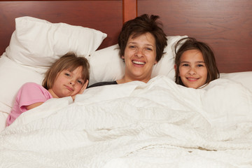 Mother and her two daughters waking up in bed with white linens. Single mother lifestyle concept.