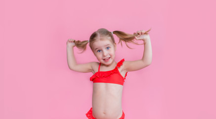 Beautiful little blond girl with two ponytales in a red swimsuit on a pink backgroung. The concept of summer family vacations, rest on the sea.