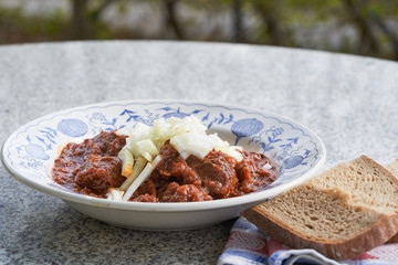 Traditional czech style spicy beef goulash, served outdoor in vintage deep plate or bowl with chopped onions on the top and rustic style bread. Food typical for eastern europe or baklan countries.