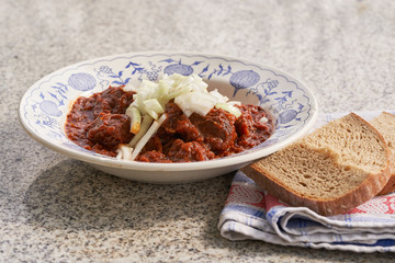 Traditional beef goulash with chopped white onion and slices of rustic bread served in vintage deep plate on the stone table outside. Popular meal in Czech republic, Slovakia, Hungary and Austria.