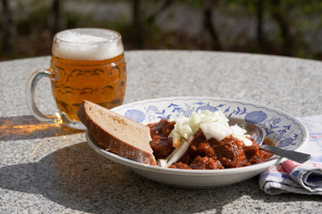 Czech style beef goulash served in deep rustic plate with slice of bread and mug with czech lager beer, on the top of the meal is chopped white onion. Food is served outside in the garden restaurant.