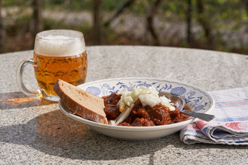 Traditional beef goulash with chopped white onion and mug of czech pilsner beer served in vintage deep plate on the stone table outside. Popular meal in Czech republic, Slovakia, Hungary and Austria.