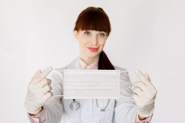 Naklejka premium Young female doctor with a stethoscope, in white coat, smiling and showing to camera protective face mask against virus infections, coved-19, standing on white background. Focus on mask in hands