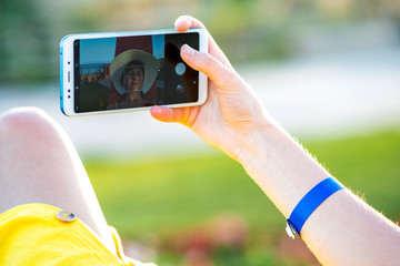 Young woman in yellow summer dress taking selfie with her mobile phone resting on a bench in park.