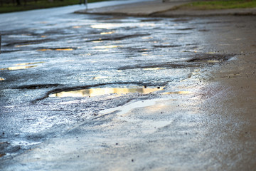 Close up of a road in very bad condition with big potholes full of dirty rain water pools.