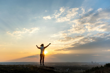 Silhouette of a woman hiker standing alone raising hands enjoying sunset outdoors. Female tourist on rural field in evening nature. Tourism, traveling and healthy lifestyle concept.