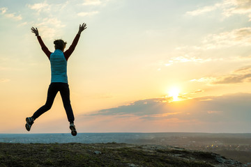 Fototapeta premium Silhouette of a woman hiker jumping alone on empty field at sunset in mountains. Female tourist raising her hands up in evening nature. Tourism, traveling and healthy lifestyle concept.
