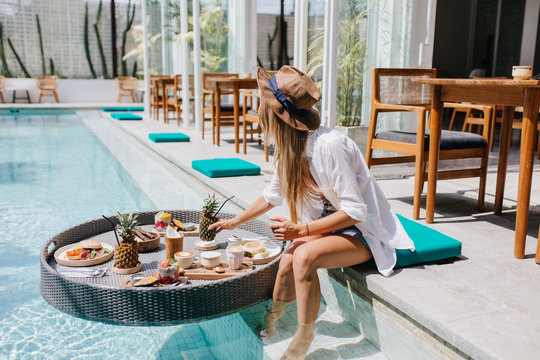 Slim Girl In Elegant Brown Hat Eating Juicy Fruits At Resort Cafe. Graceful European Woman In White Shirt Relaxing With Cocktail And Food In Pool.