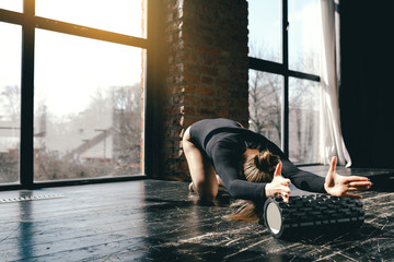 Flexible gymnast is sitting on the floor and doing forward bends on massage roller. The yogi bends...