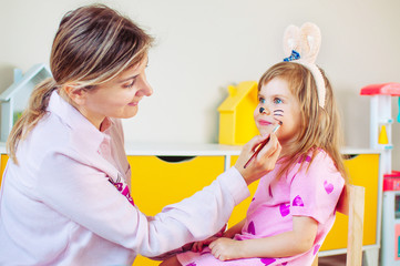 Mother painting face of her daughter as a bunny in the kids room