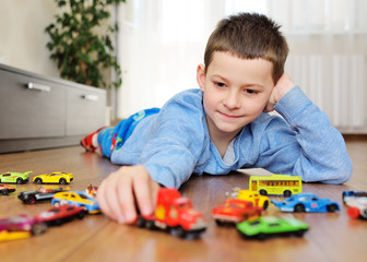  a preschool boy in a blue sweater is lying on the wooden floor playing with toy cars against the...
