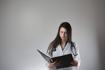 Fototapeta premium Female healthcare professional examining patient chart isolated on grey background. Stethoscope around neck, white coat on and buttoned.