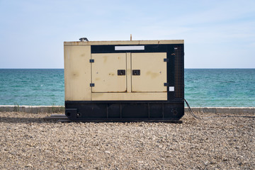 An electric diesel generator stands on the beach against the background of the sea and blue sky. An...