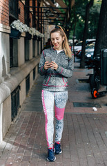 Happy female walks through the city before running looking at her smartphone and listening to music
