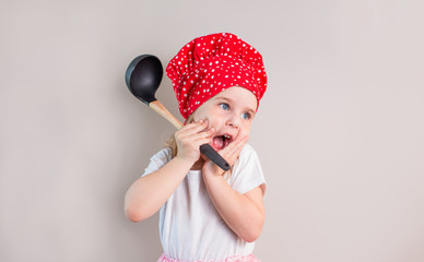 Surprised little blonde girl in red cook hat with ladle on a neutral grey background, copyspace