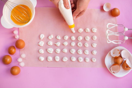 The process of making meringue cookies step by step. In the hands of a cooking bag for cream, squeezes out the meringue. On a pink background, parchment paper for baking, eggs, hand mixer, whisk.