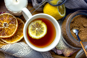Hot black tea with a slice of lemon in a large gray mug with a spoon closeup, top view, next to dried oranges, fresh lemon, brown sugar, gray napkin on a wooden tray. The concept of a cozy home tea 
