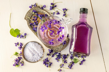 Obraz na płótnie Canvas Cup of herbal violet violetta odorata tea with fresh spring blossom petals flowers on white wooden background with bottle of sirup, tincture, sugar, salt, body oil