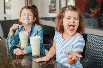 Two funny Caucasian little preschool sisters siblings drink milk shakes in a cafe. Friends girls having fun together. Cold summer desserts for kids. Happy authentic childhood lifestyle.