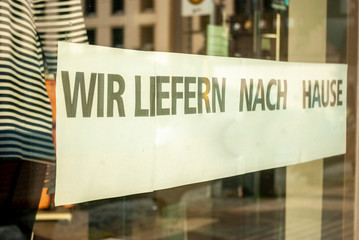 Schaufenster von Ladengeschäft mit Schild „Wir liefern nach Hause“, in Deutsch, mit Auslagen und die Spiegelung der Umgebung in Unschärfe zur Zeit der Corona Covid-19 Pandemie im März 2020