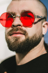 Closeup portrait of a bearded man with a shiny earring in his ear. Stylish attractive guy in black is looking at the camera. Serious harsh guy in red stylish sunglasses. Brutal handsome guy.