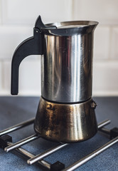 Close-up on an old, traditional Italian coffee maker placed on a metal pot stand on blue kitchen counter top.