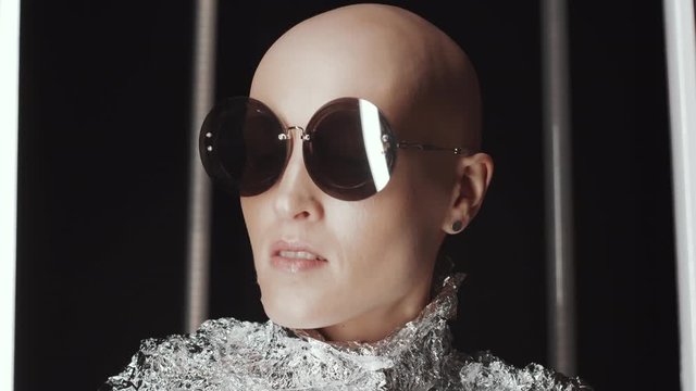 Chest up shot of young bald woman in trendy sunglasses and foil wear looking around while posing for camera in dark studio with flickering fluorescent lamps