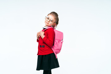 Adorable little girl in red school jacket, black dress, rounded glasses holding on to the straps of a backpack and smiling and looking at the camera, posing on white studio background. Isolate.