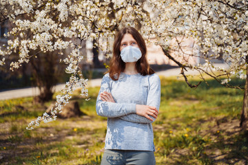 Spring and coronavirus. A European girl in a home-made protective face mask standing in a park lit by the sun against a background of blooming trees. Virus covid-19 protected.