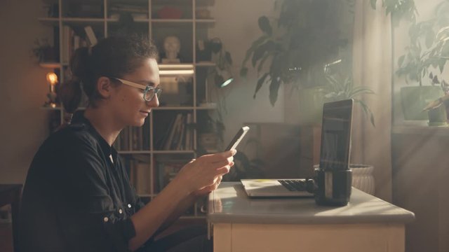 Girl freelancer works at a laptop at home. She receives a notification on the phone, she is happy with the message, puts off the phone and continues to work. Sun glare from the window. Against the