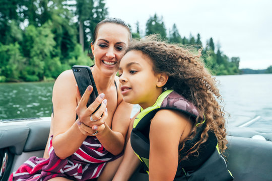 Smiling mom showing daughter video on smart phone on boat on lake surrounded by trees