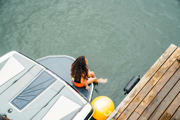 Girl in life jacket sitting on boat next to dock on lake with feet in water