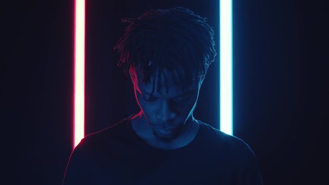 Chest up shot of young African American man raising his head and looking at camera while posing in dark studio with flickering neon light of blue and red colors