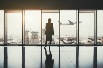 A silhouette of a successful woman entrepreneur standing indoors of a luxurious interior with a reflective marble floor, and a plane gaining altitude over an urban skyline outside a panoramic window