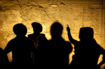 Dark abstract view of unrecognizable silhouettes in front of glowing wall of hieroglyphs; selective...