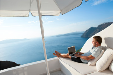 Man relaxing with a laptop computer on white balcony with bright scenic view of the Mediterranean Sea and Santorini caldera