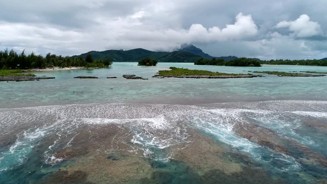 Beautiful Drone shot of lush tropical landscape flying towards the lagoon reef break of vacation paradise Bora Bora, French Polynesia. Moody rain clouds fill the South Pacific sky