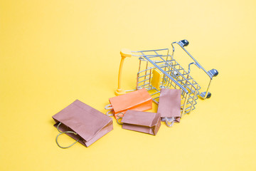 Fototapeta na wymiar overturned miniature shopping trolley on a yellow background, fallen small paper bags, copy space, online shopping concept